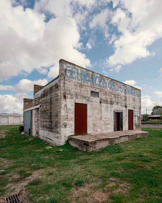 Weathered white concrete ice house with faded blue Celina Ice & Cold Storage Co. lettering, rust-red doors, and a wide cloudy sky
