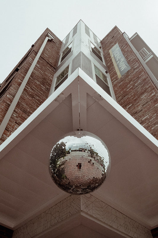 A large mirrored disco ball hanging beneath the sharp corner of a brick-and-metal building, seen from below
