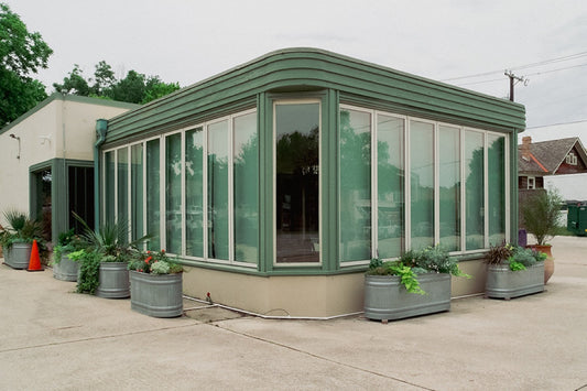 A rounded green-trimmed building wrapped in tall glass windows, with galvanized planter tubs of greenery out front