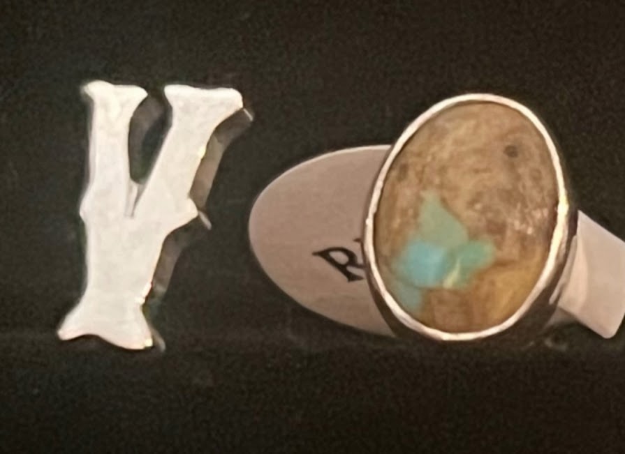 Turquoise initial ring — close view, silver initial and brown-matrix oval stone with a blue patch, paper tag behind, on black