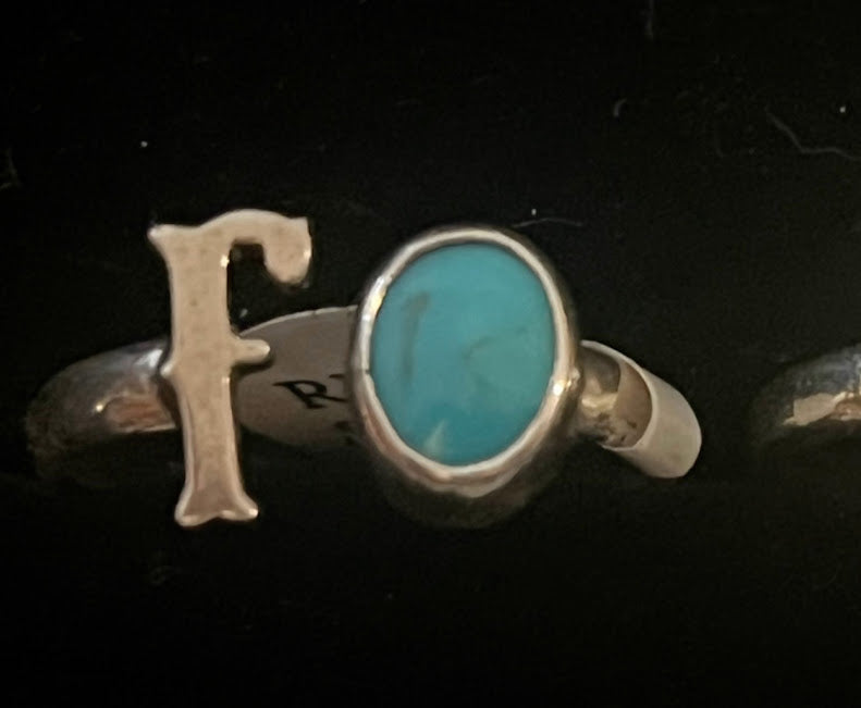 Turquoise initial ring — letter F in sterling silver beside an oval blue stone, paper tag on the band, on black