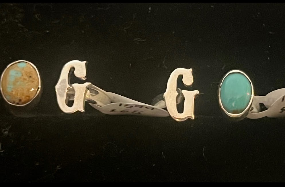 Turquoise initial ring — two letter G rings side by side, oval stones, paper price tags on the bands, on black