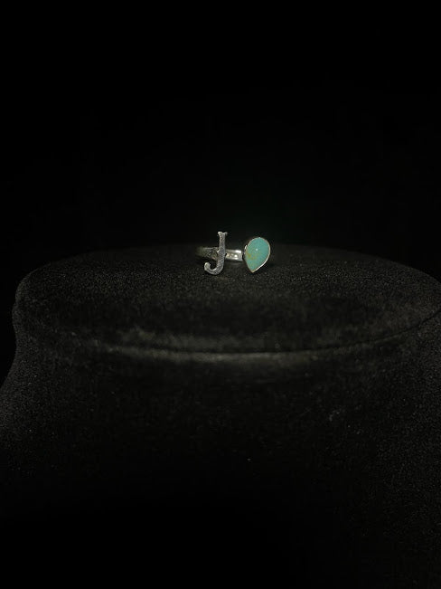 Turquoise initial ring — letter J in sterling silver beside a pale green teardrop stone, on black velvet