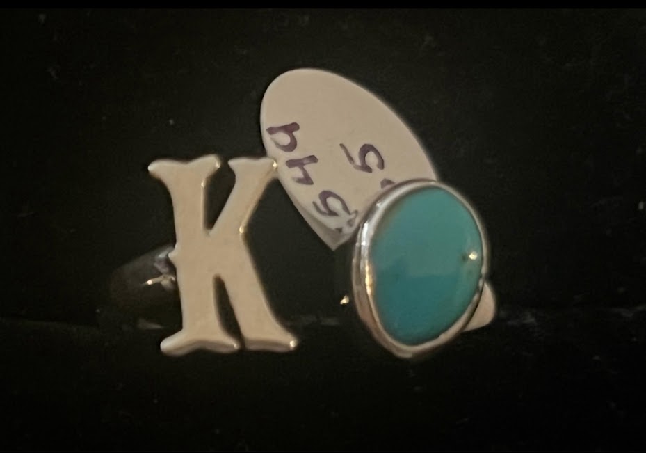 Turquoise initial ring — letter K in sterling silver beside an oval blue stone, paper price tag behind, on black
