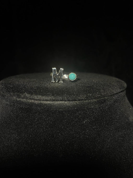 Turquoise initial ring — letter M in sterling silver beside a small round blue stone, on black velvet