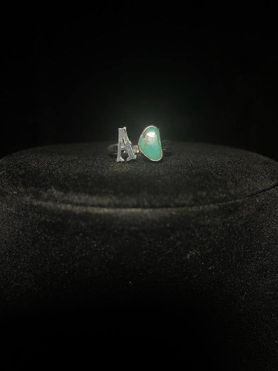 Turquoise initial ring — letter A in sterling silver beside a green teardrop stone, on black velvet