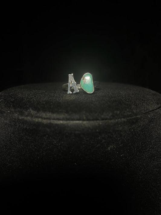 Turquoise initial ring — letter A in sterling silver beside a green teardrop stone, on black velvet